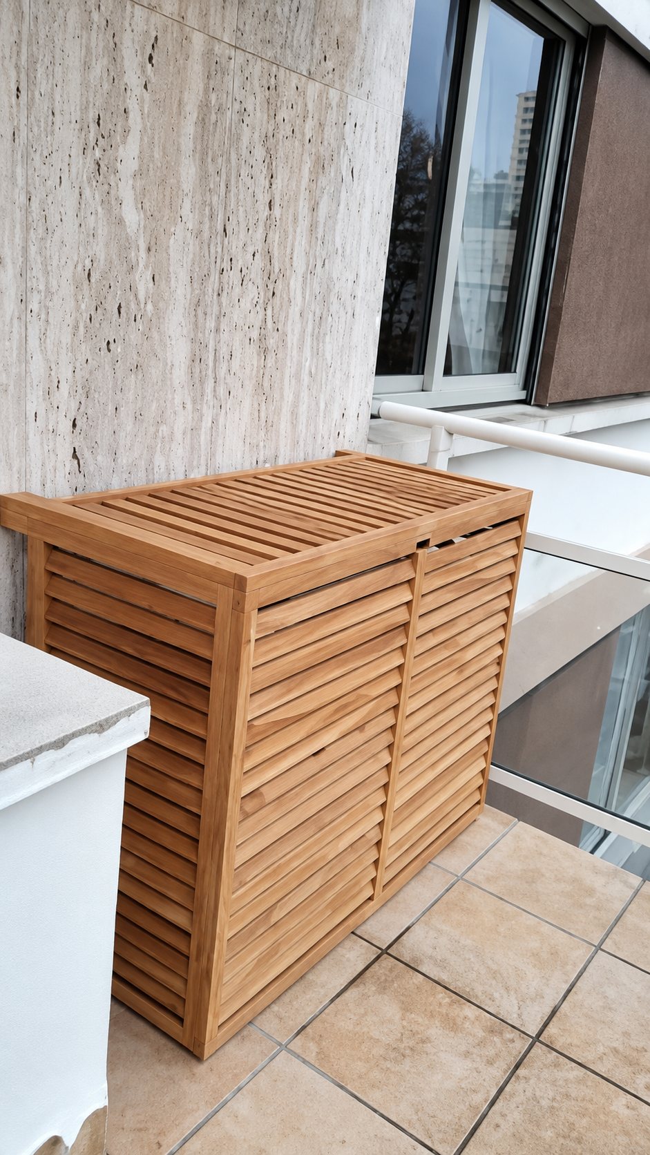 Un cache en bois à lamelles, destiné à masquer un climatiseur, est installé sur une terrasse carrelée devant un mur en pierre claire.