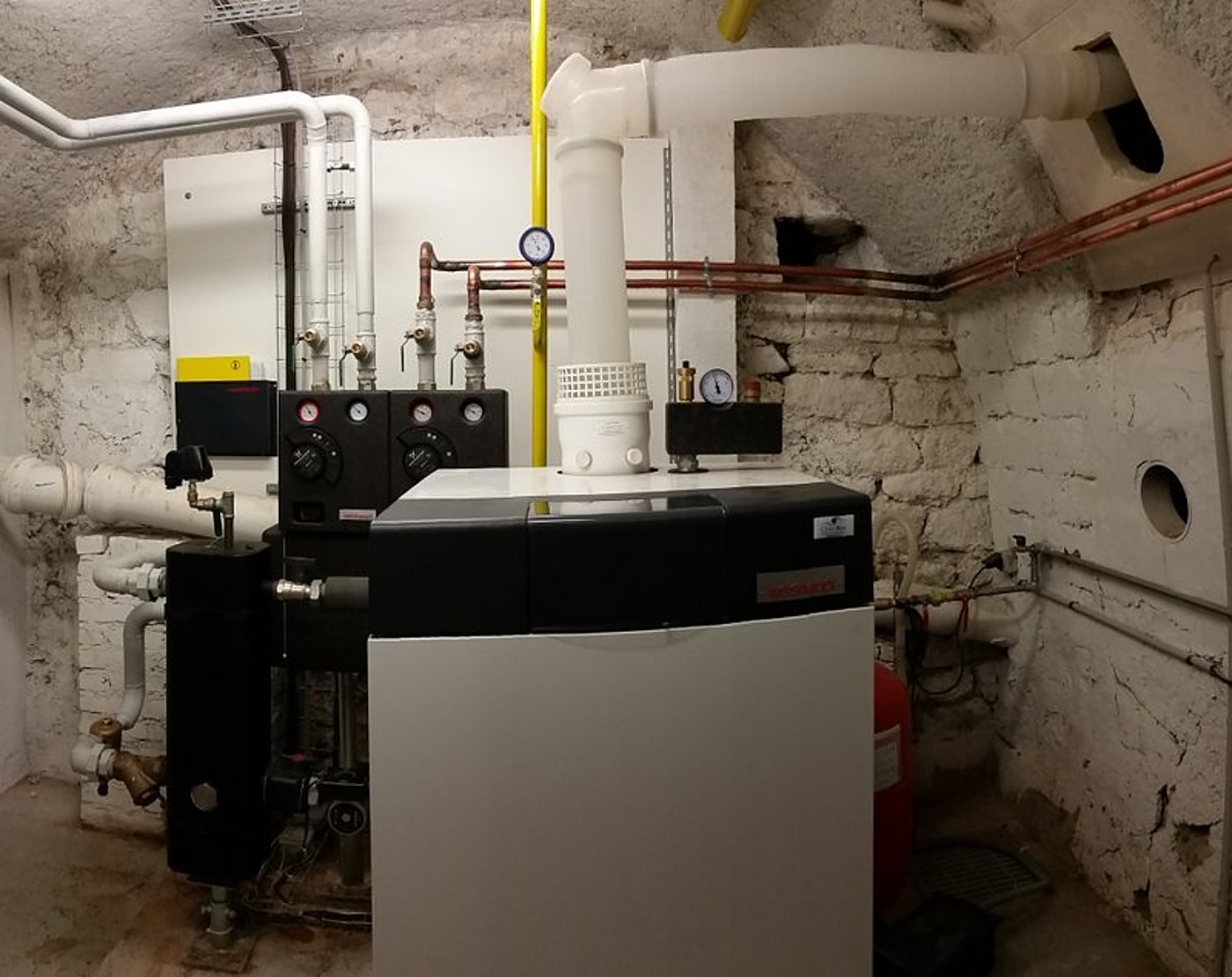 Une chaudière à gaz blanche et noire avec ses tuyaux en cuivre et ses conduits blancs est installée dans un local technique aux murs de pierre blanchis.