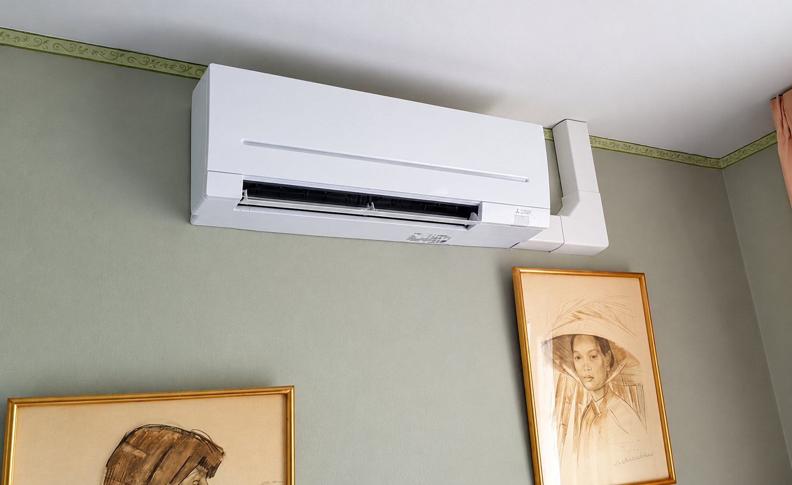 Une unité intérieure de climatisation blanche est fixée en haut d'un mur vert sauge, avec un conduit blanc sur le côté et deux tableaux encadrés en dessous.