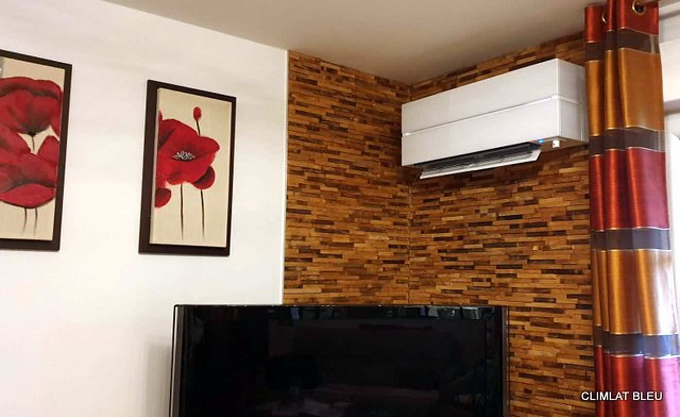 Un climatiseur mural blanc est fixé dans l'angle d'un mur en lattes de bois, près d'une télévision, de deux tableaux de coquelicots rouges et d'un rideau rayé.