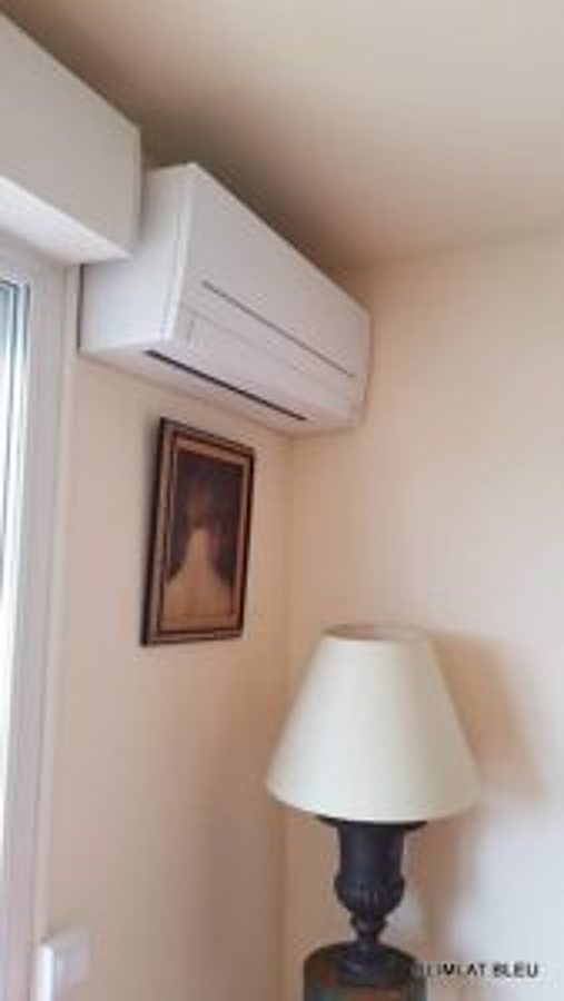 Un climatiseur mural blanc est fixé en haut d'un mur beige, à côté d'un tableau encadré et d'une lampe à abat-jour clair.