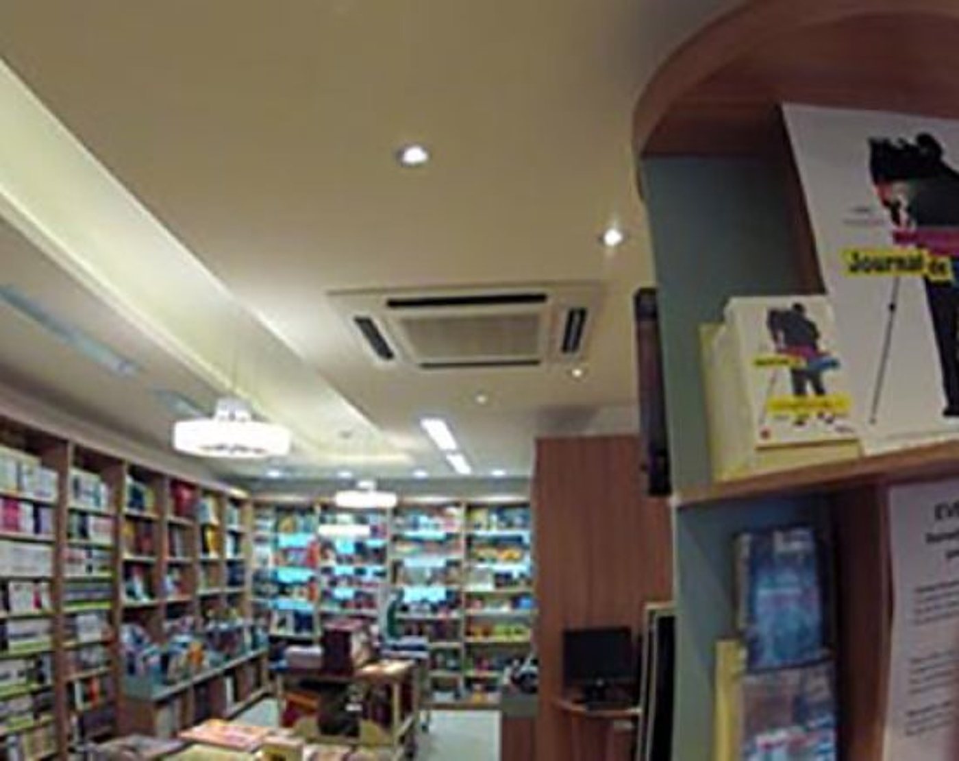 Intérieur d'une librairie aux longues étagères en bois remplies de livres, avec un présentoir au premier plan et un plafond équipé de spots et d'une climatisation.