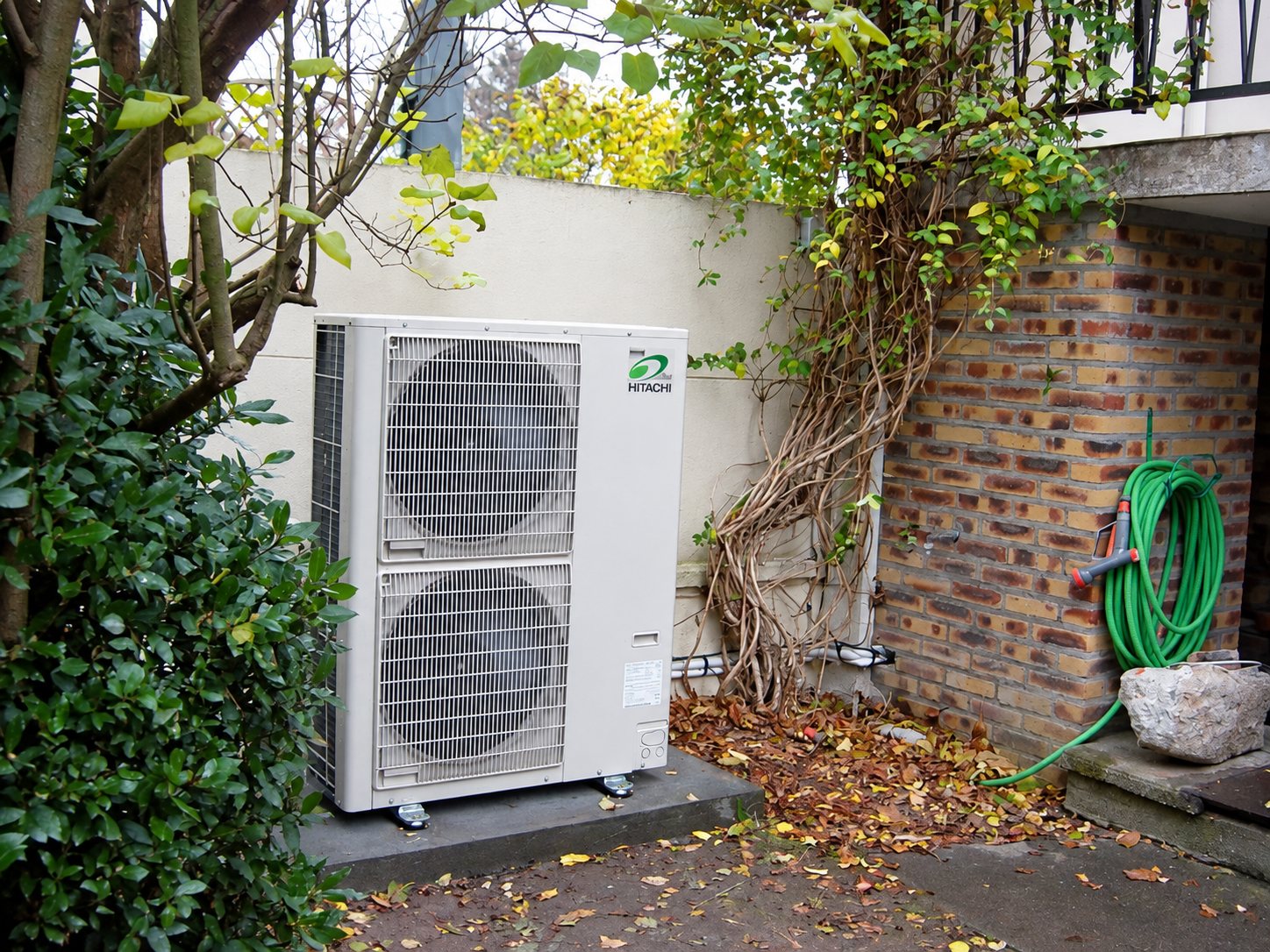 Une unité extérieure de pompe à chaleur Hitachi posée sur une dalle dans un coin de jardin, entre un mur blanc, un mur de briques et de la végétation.