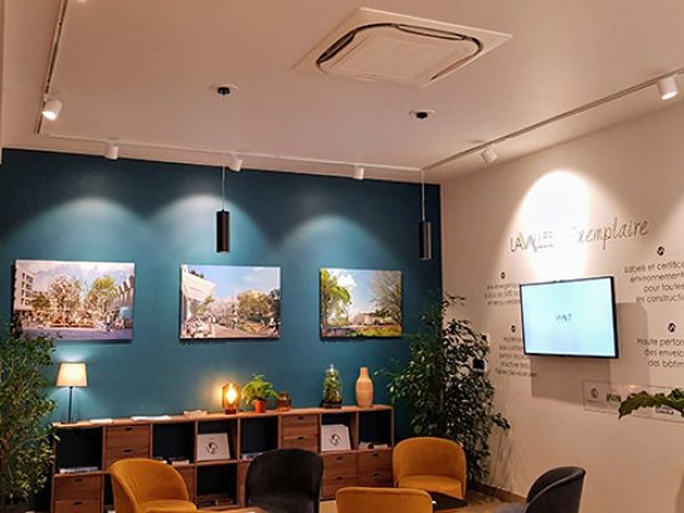 Intérieur d'un espace d'accueil avec un mur bleu canard orné de trois visuels de projets, un meuble bibliothèque en bois, des fauteuils jaunes et un écran mural.