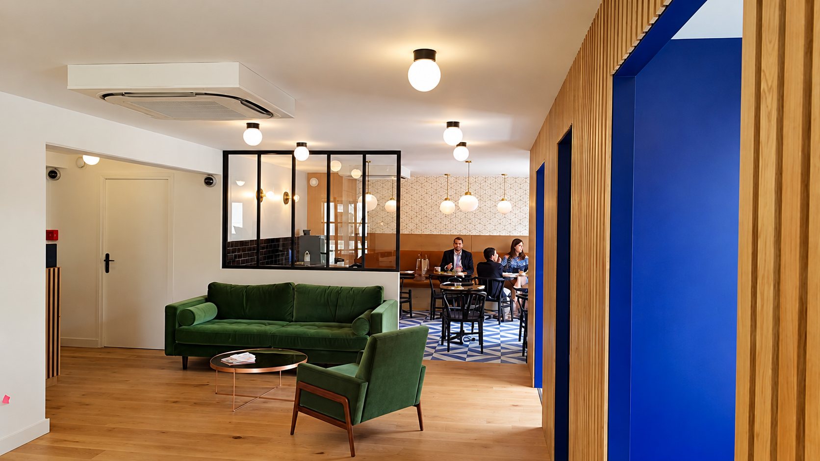 Un intérieur moderne avec un canapé et un fauteuil en velours vert, des lamelles de bois et des cadres bleus, et au fond des personnes attablées derrière une verrière.